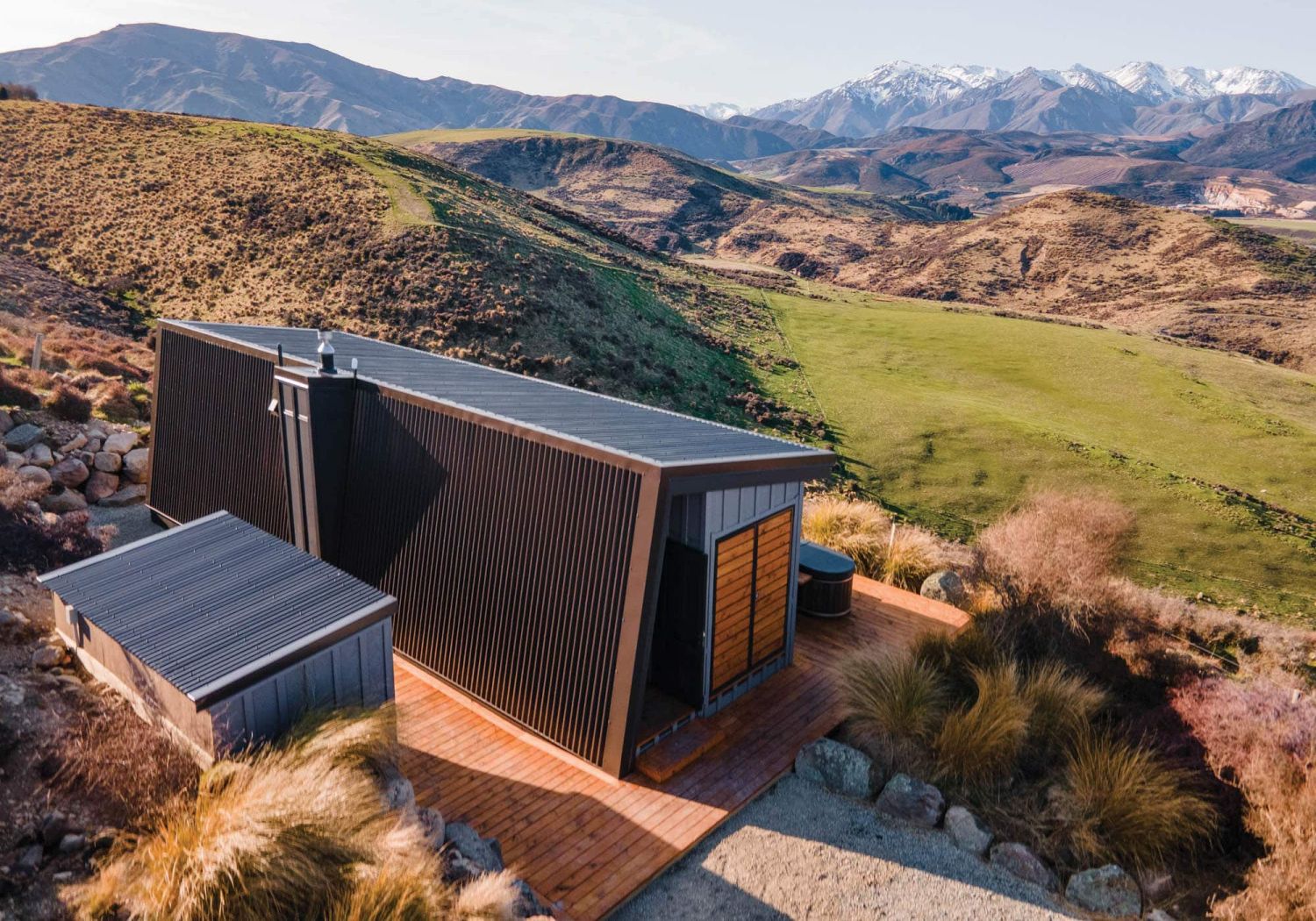 Compact black-clad high-country cabin with timber deck set among tussock, snow-capped ranges behind – site-responsive design by Chilton + Mayne