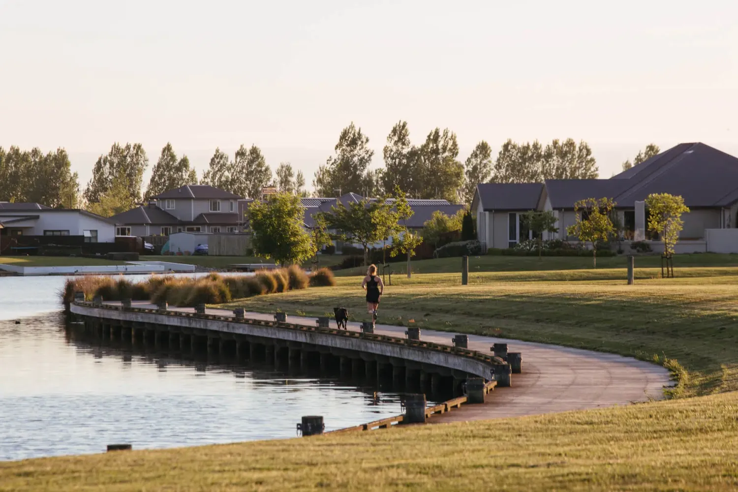 Lakeside boardwalk and homes in a Canterbury subdivision at dusk, with a runner and dog on the path